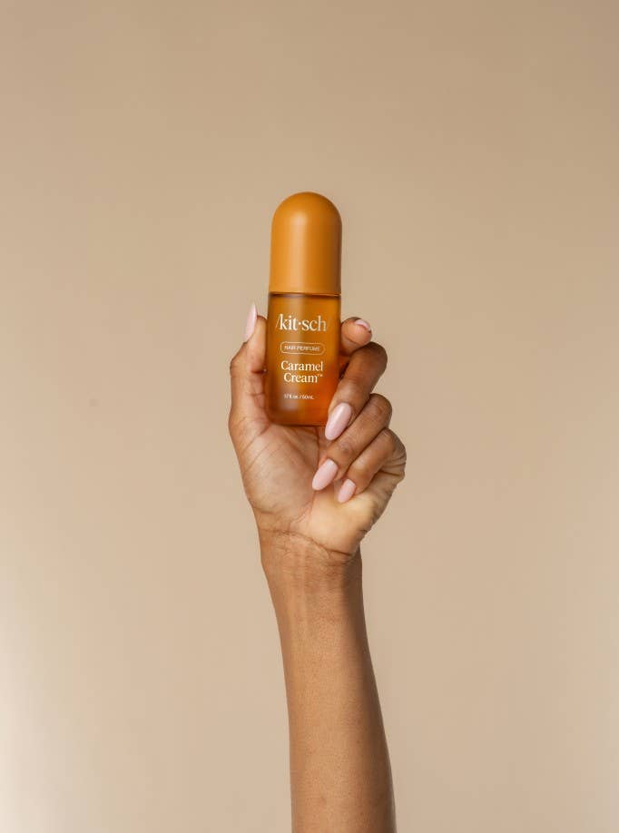A hand with light pink manicured nails holds a small amber bottle of kitsch Caramel Gloss in front of a solid beige background.