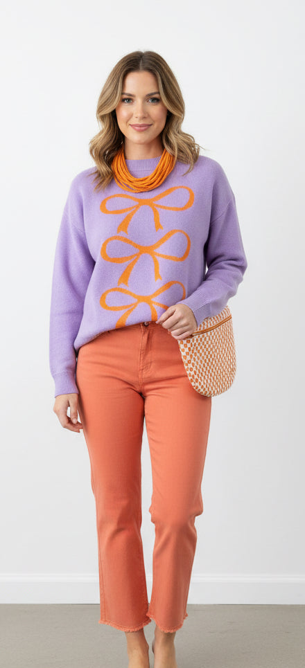 Woman wearing a purple sweater with orange bow design, orange pants, and an orange scarf against a white background