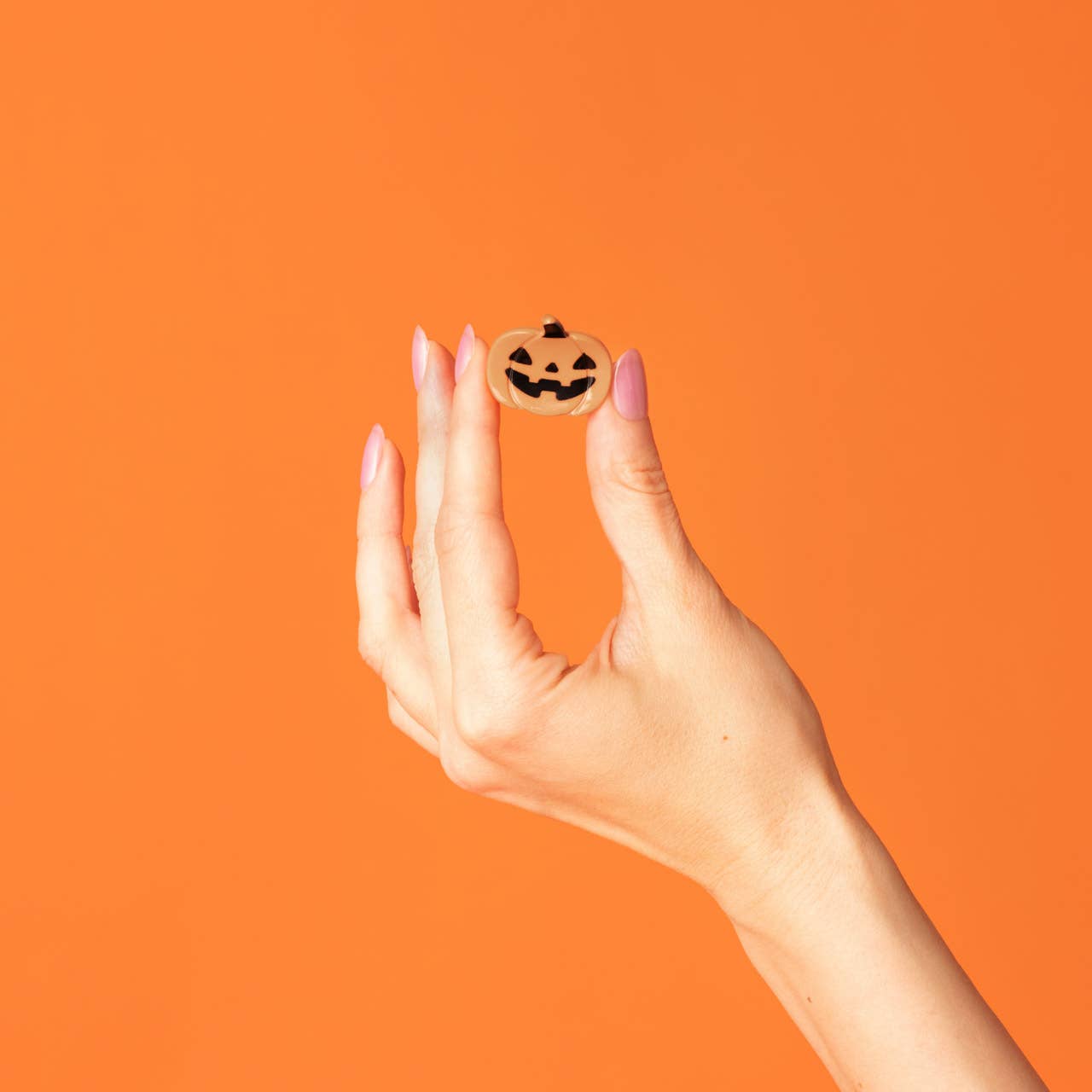 A hand with pink manicured nails holds a small round wooden pumpkin charm with a carved jack-o'-lantern face against a solid orange background.