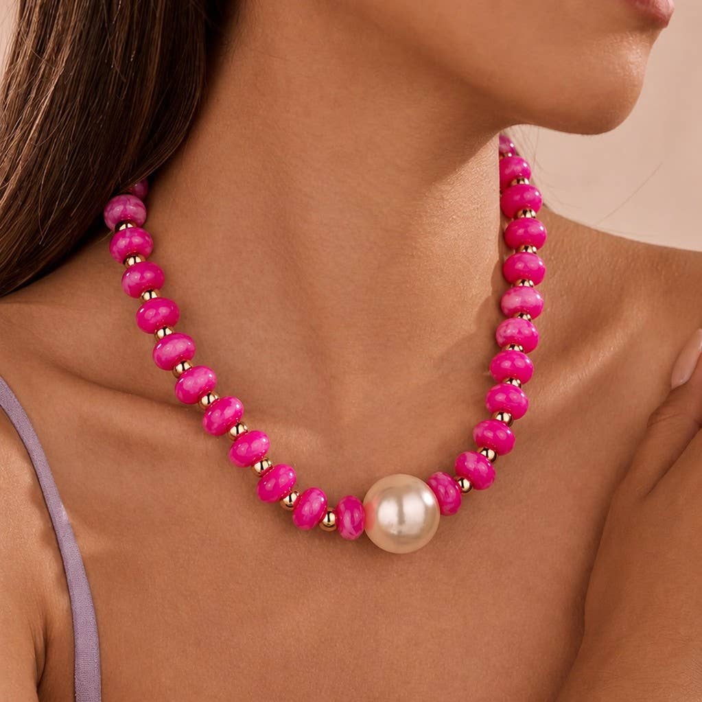 A close-up of a woman wearing a vibrant pink beaded necklace with a large white pearl at the center, set against a soft beige background.