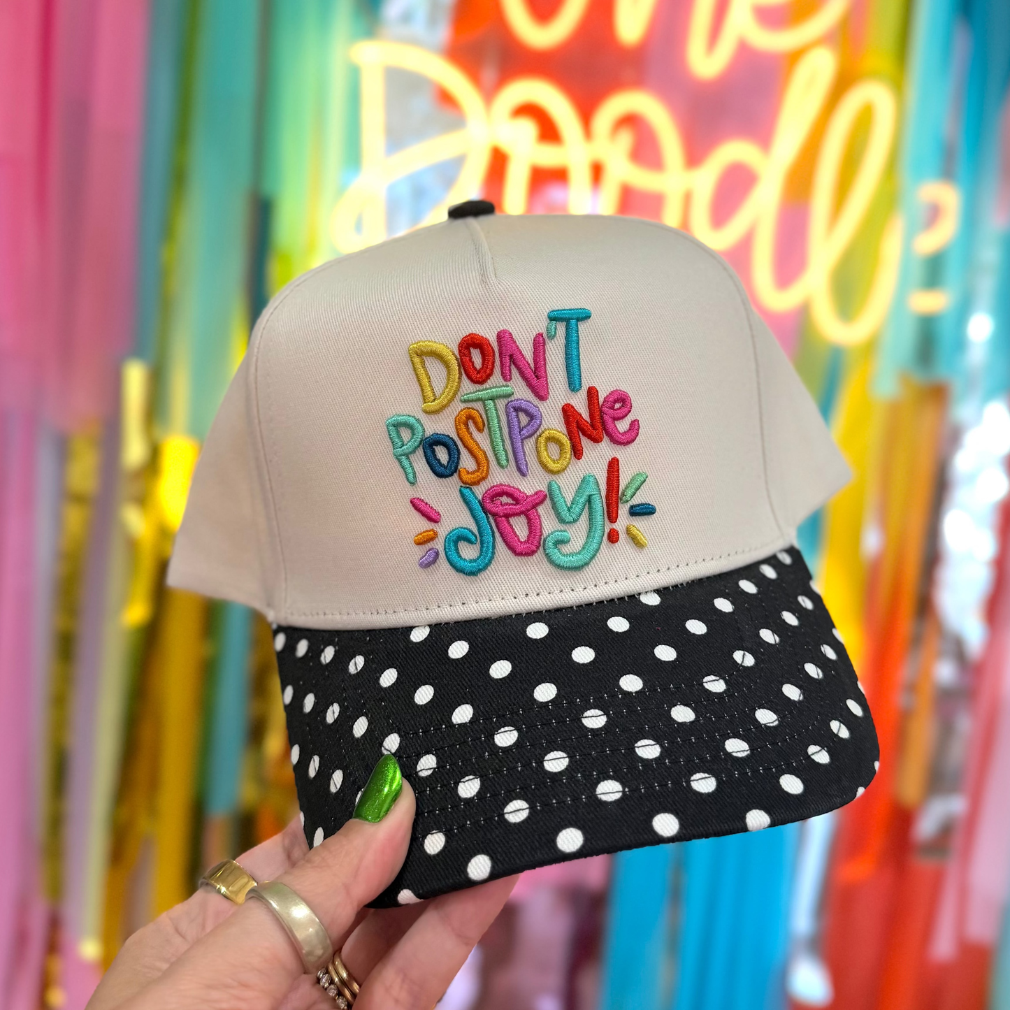 Cap with colorful text 'DON'T POSTPONE JOY!' held in front of a vibrant, multicolored background.
