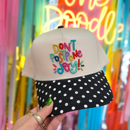 Cap with colorful text 'DON'T POSTPONE JOY!' held in front of a vibrant, multicolored background.