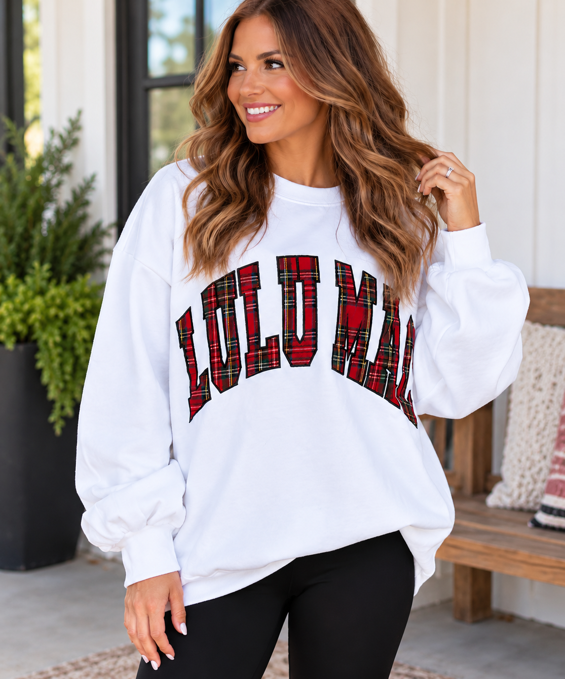 Woman wearing a white Lulu Mac oversized crewneck sweatshirt with bold red and black plaid lettering, styled with black leggings.