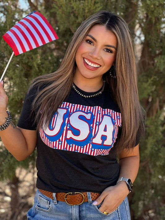 Woman wearing a black t-shirt with 'USA' print, holding an American flag outdoors.
