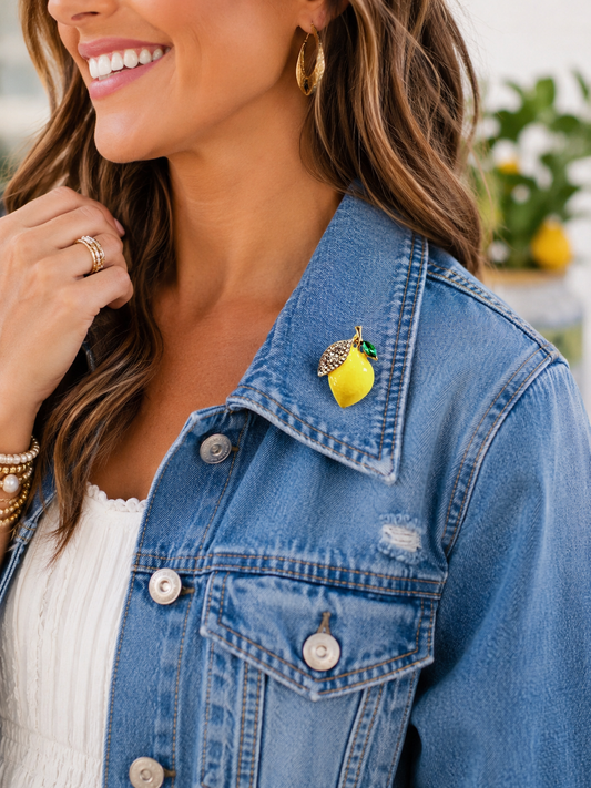 Woman wearing a denim jacket with a lemon-themed brooch, smiling outdoors.