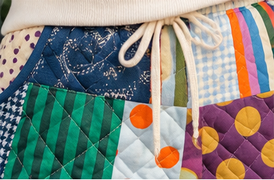 Close-up of a colorful patchwork quilted fabric item with a cream drawstring, showing multiple patterned panels including green stripes, orange polka dots, purple polka dots, blue gingham, and striped sections.