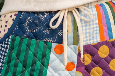 Close-up of a quilted patchwork fabric item, likely a bag or pouch, featuring a cream drawstring closure and multiple colorful patterned panels including stripes, polka dots, gingham, and solid fabrics.