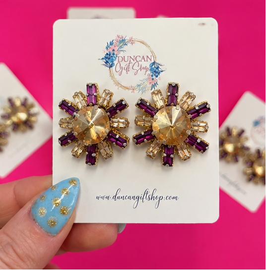 A collage showing a pair of floral cluster stud earrings on display cards, held by a hand with blue patterned nail polish against a bright pink background. The earrings feature gold-tone metal with faceted clear crystals and dark purple crystals.