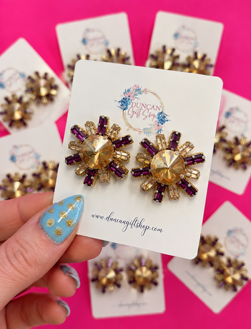 A hand with blue and gold nail art holds a white display card featuring a pair of gold and purple starburst stud earrings. The background is a bright pink surface with several blurred copies of the same earring cards.