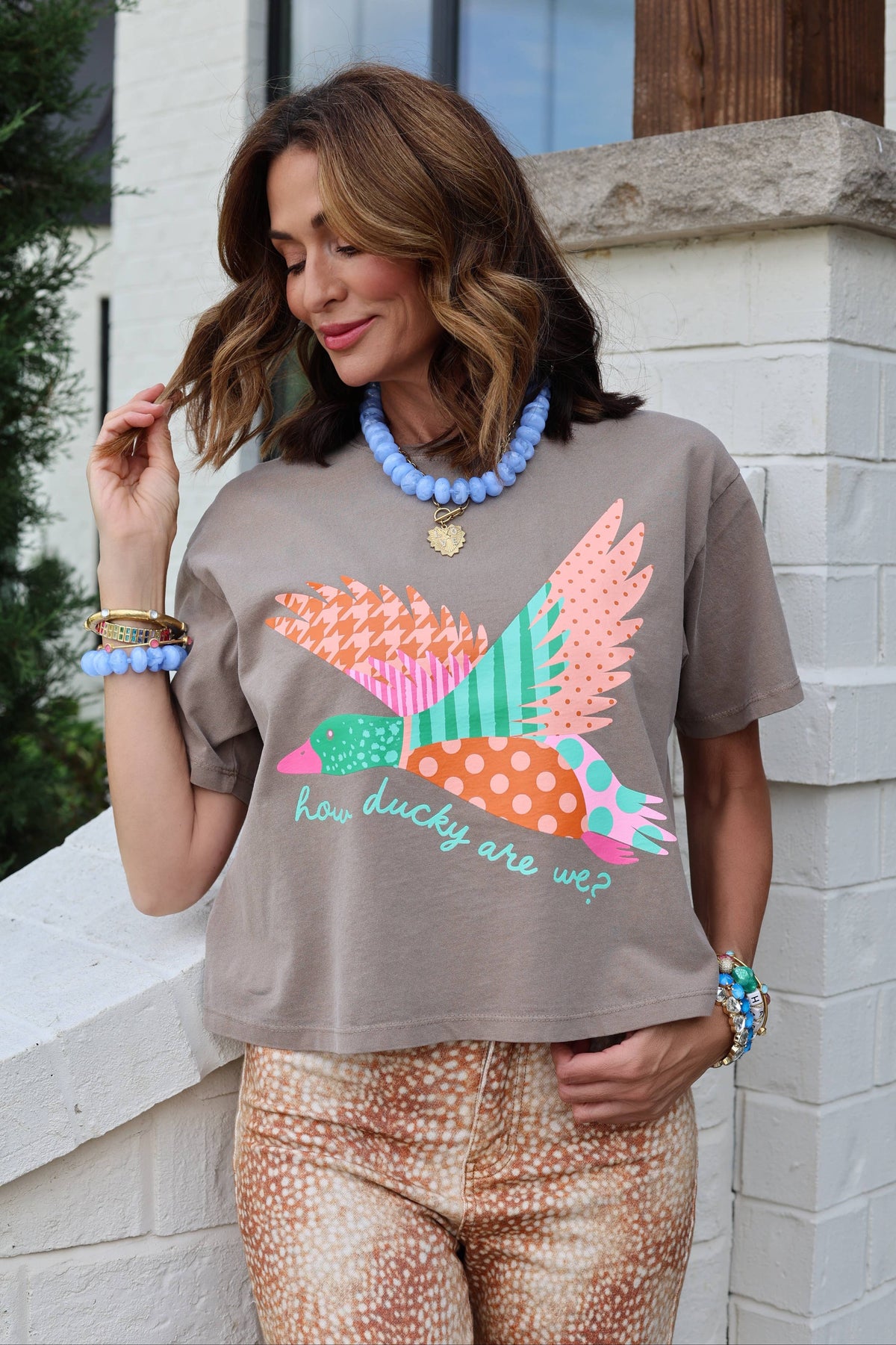 A woman wearing a poppy & pone taupe graphic t-shirt with a colorful bird design and the text 'how lucky are we?', paired with patterned pants, standing outdoors against a white brick wall.