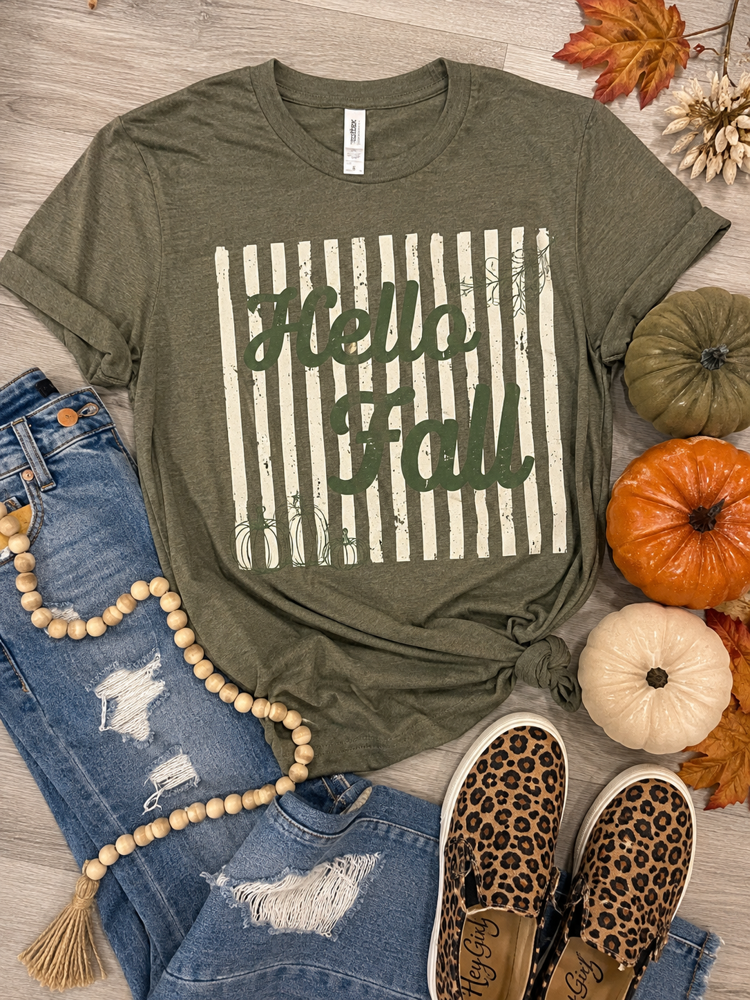 An olive green graphic t-shirt with 'Hello Fall' printed in green script over a cream and green striped background, styled in a flat lay with blue jeans, leopard print slip-on shoes, wooden beads, and small pumpkins on a light wood floor.