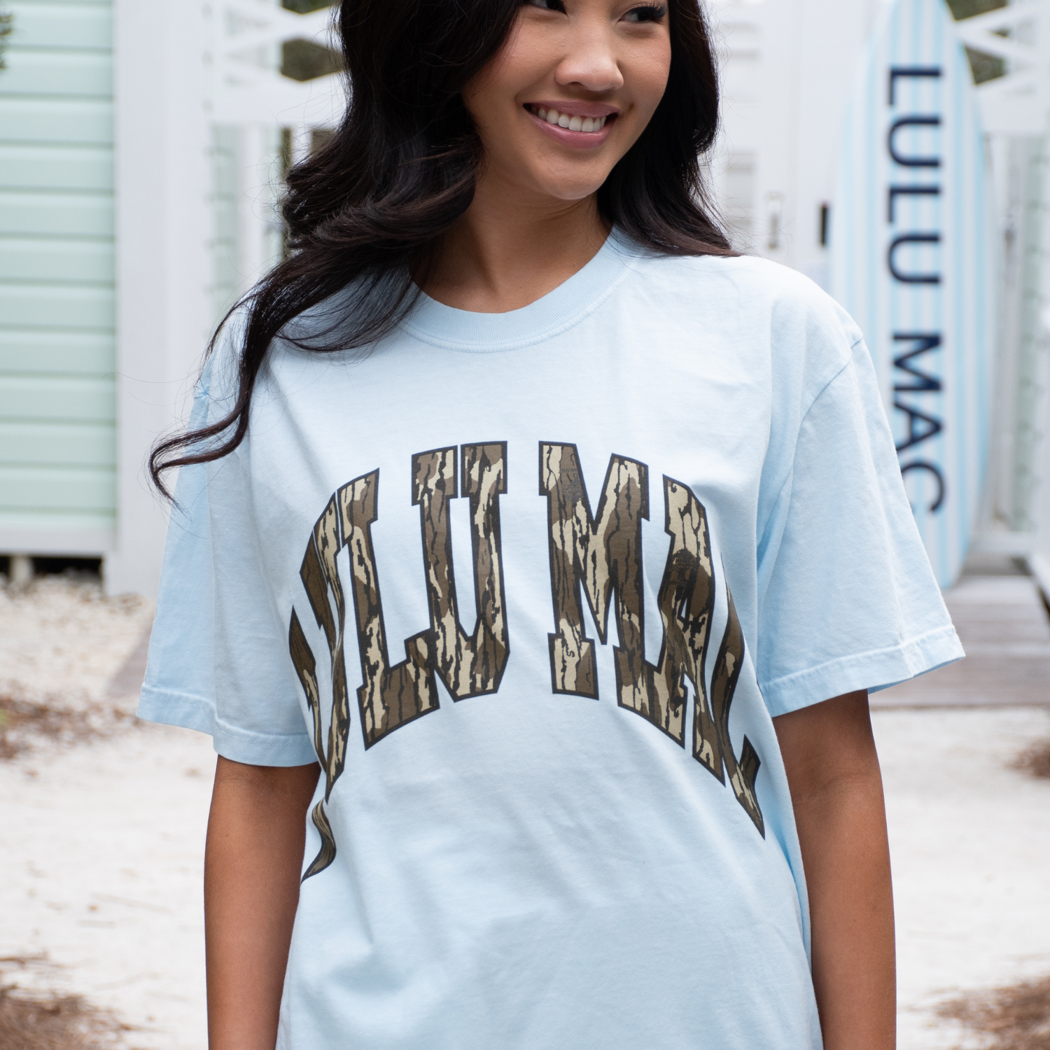 Woman wearing a light blue t-shirt with camo 'LuLu Mac' printed on it, standing outdoors.