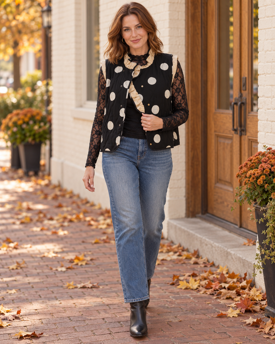 A woman walking outdoors on a brick sidewalk in front of a white brick building, wearing a black long-sleeve vest with beige and white polka dots over a black top, blue jeans, and black ankle boots.