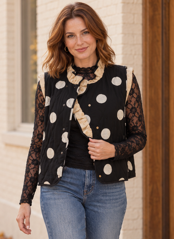 A woman wearing a black sleeveless quilted vest with large beige polka dots over a black lace long-sleeve top and blue jeans, standing outdoors against a light brick wall.