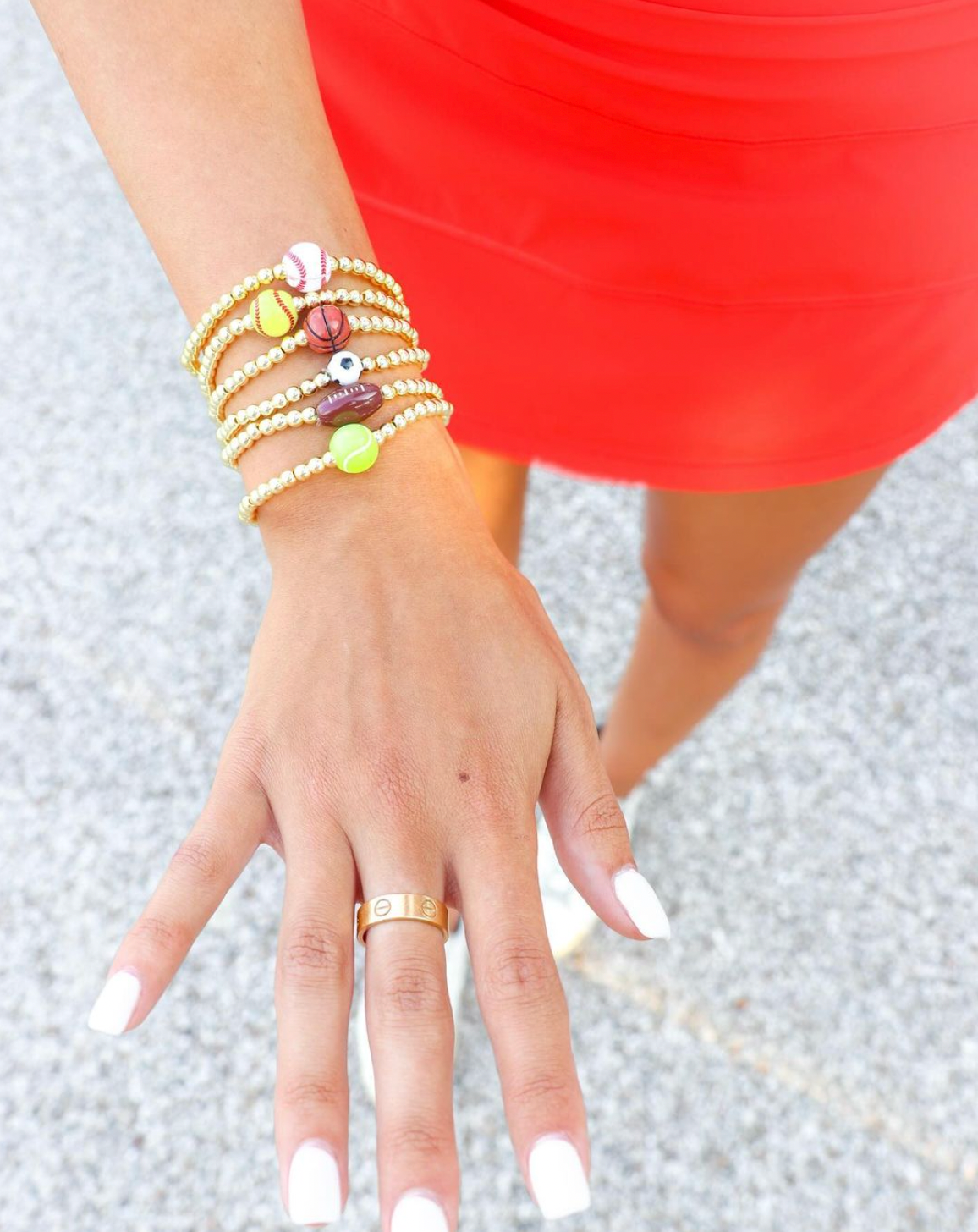 A close-up of a person's hand and wrist wearing a stack of gold beaded bracelets with colorful charms, paired with a gold ring, against a bright red dress and gray pavement background.