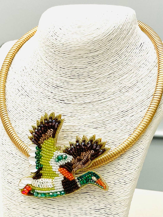 A gold-tone beaded necklace displayed on a white textured mannequin bust. The necklace features a prominent front pendant shaped like a flying duck or goose, decorated with green, white, black, brown, and orange beads.
