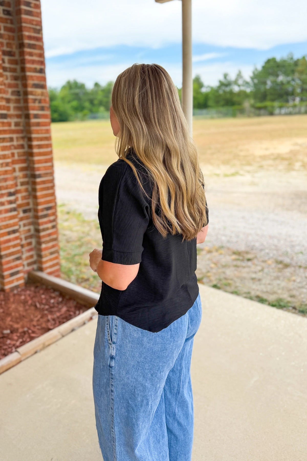 Person wearing a zenana black shirt and blue jeans standing near a brick wall with a scenic background.