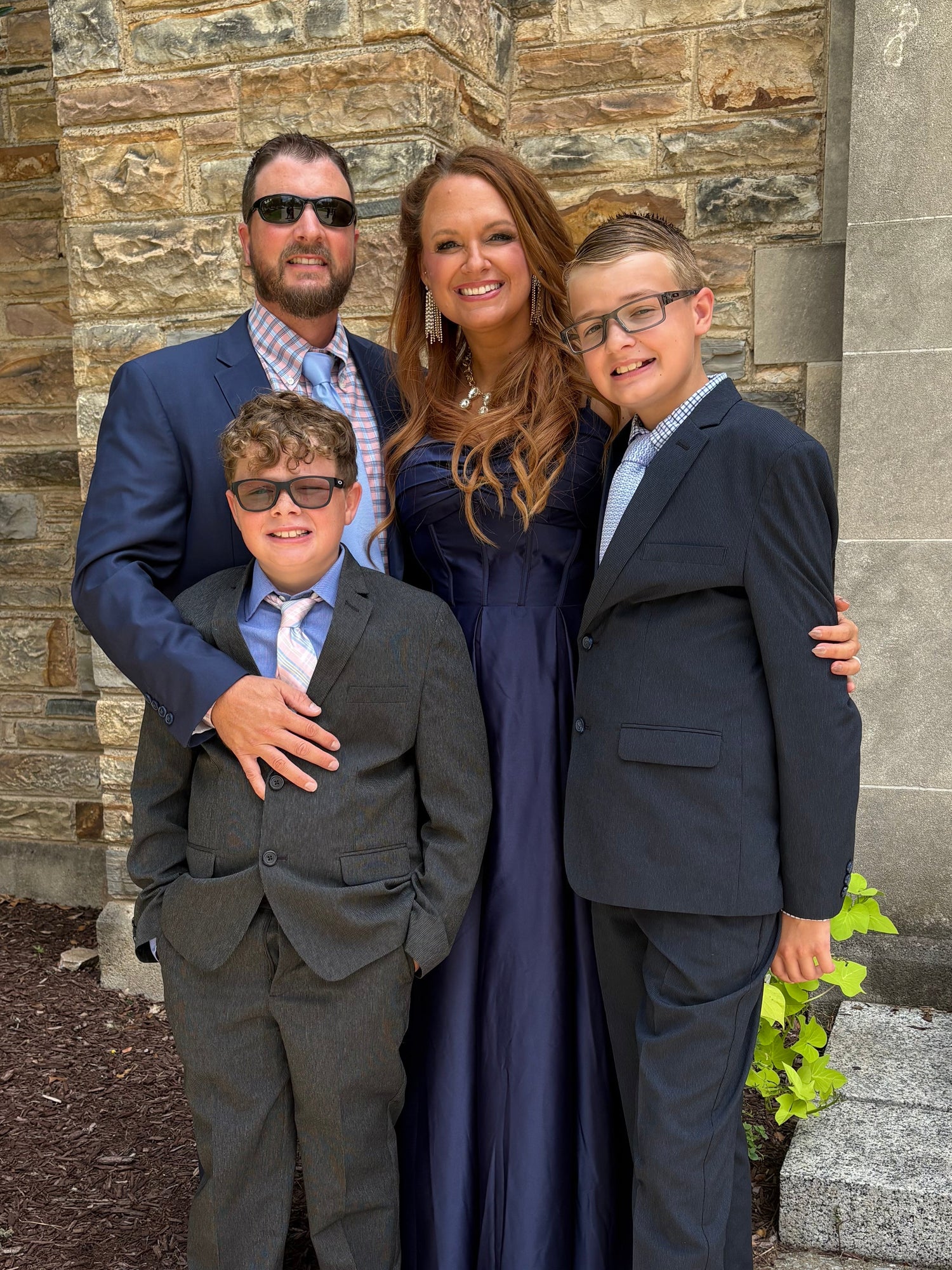A family of four posing outdoors in front of a stone building. The mother wears a long navy blue gown, while the three sons wear formal suits in gray and navy blue.