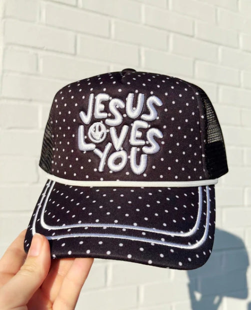 Black and white polka dot cap with 'Jesus Loves You' text held against a white brick wall.