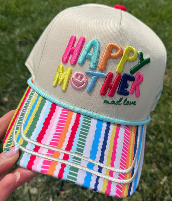 A hand holds a baseball cap outdoors on grass. The cap has a light beige crown with colorful embroidered text reading 'HAPPY MOTHER mad love' on the front, and a bright multicolor striped brim.