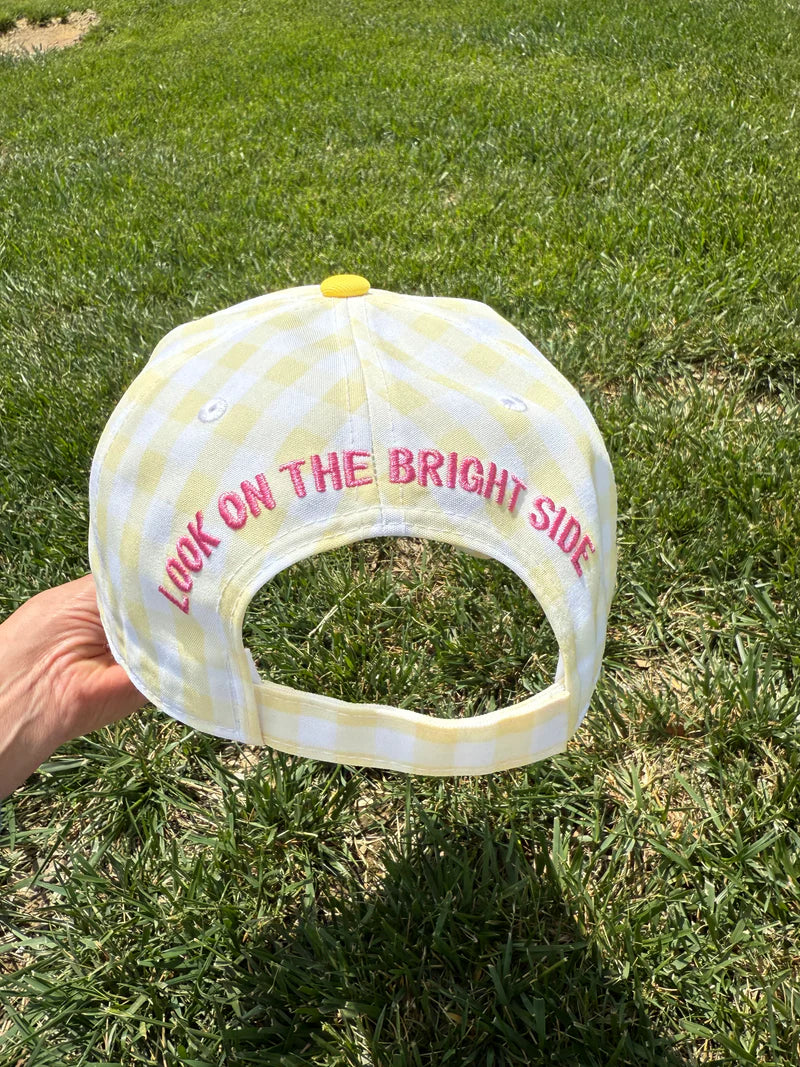 A light yellow and white gingham baseball cap held by a hand against a green grass background. The cap features pink embroidered text reading 'LOOK ON THE BRIGHT SIDE' and a small yellow button on top.