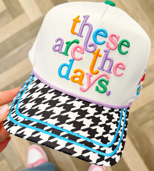 A white snapback cap held in a hand, featuring colorful embroidered text reading 'these are the days.' on the front, a black-and-white houndstooth patterned brim, and light purple piping around the edge of the brim.