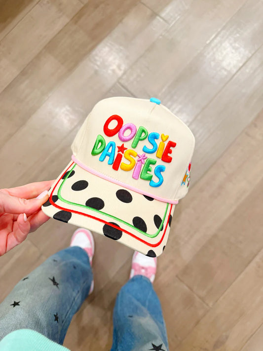 Cap with 'Oopsie Daissies' text held by a person on a wooden floor.
