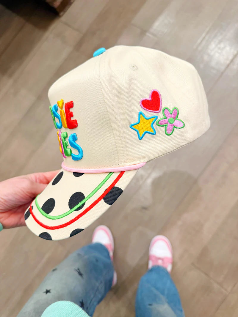 Beige cap with colorful text and designs held by a person on a wooden floor.