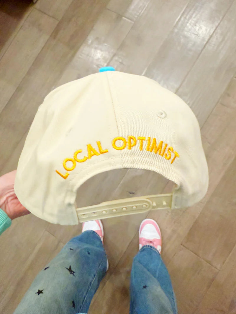 Beige cap with 'LOCAL OPTIMIST' text held by a person on a wooden floor.