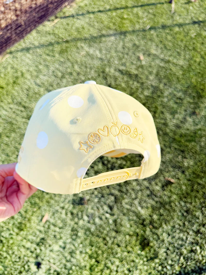 Yellow cap with white polka dots and text held in front of a grassy background