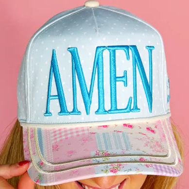 A close-up of a snapback cap being held by a person, featuring a light blue dotted crown with large blue embroidered 'AMEN' lettering on the front, and a pink floral visor with a gingham brim.