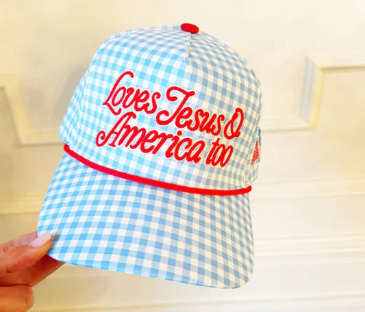 Blue and white checkered cap with red text 'Loves Jesus & America too' held by a hand.