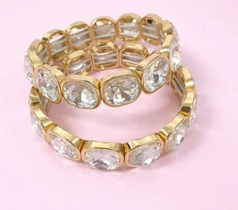 A stack of two gold-plated stretch bracelets, each set with clear oval rhinestones, displayed on a solid light pink background.
