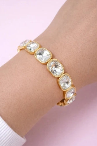 A gold-toned cuff bracelet featuring five large, clear cushion-cut rhinestones set in a gold-plated band, shown worn on a person's wrist against a soft pink background.