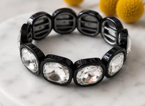 A black beaded stretch bracelet with large clear faceted oval rhinestones, displayed on a white marble surface with yellow pom-pom decorations in the background.