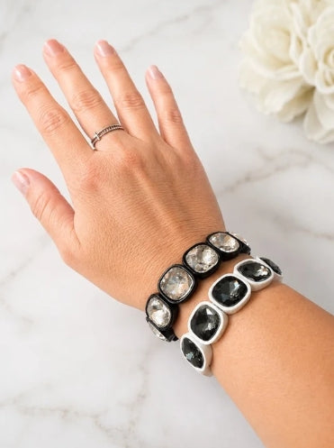 A close-up of a person's hand and wrist wearing a stack of three black and silver cuff bracelets with large clear faceted gemstones, set against a light marble background.