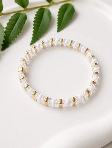 A beaded stretch bracelet with white pearl-like beads and small gold spacer rings, displayed on a white surface with green leaves in the background.