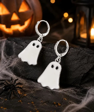 A pair of silver hoop earrings with ghost-shaped charms, displayed on a dark rock with Halloween props including a spider, cobwebs, and a glowing jack-o'-lantern in the background.