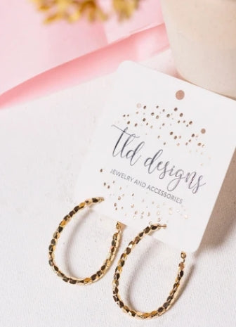 Gold textured hoop earrings displayed on a white card labeled 'lid designs jewelry and accessories' on a light surface with soft pink and beige background elements.