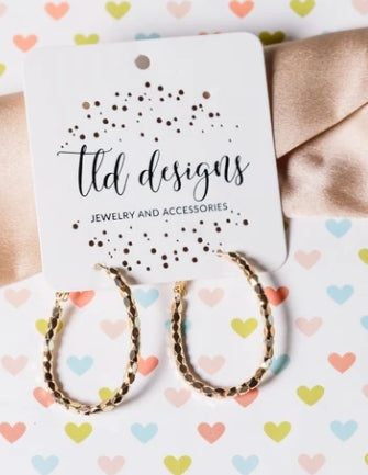 Gold hoop earrings with a braided texture, attached to a white display card printed with 'ltd designs JEWELRY AND ACCESSORIES', set against a white background with pastel heart patterns and a beige satin ribbon.