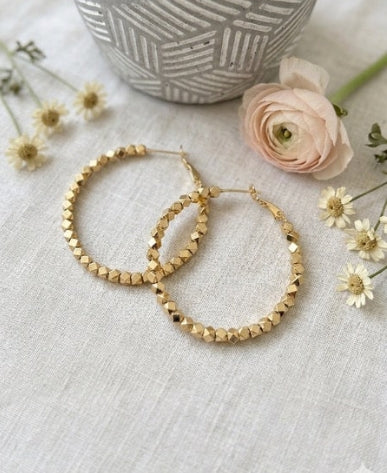 A pair of chunky gold hoop earrings with a textured, faceted beaded design, displayed on a light beige linen fabric alongside a pink rose, small white daisies, and a gray patterned ceramic pot.
