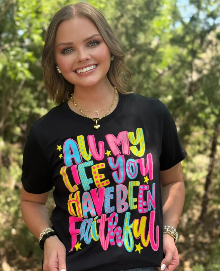 A woman smiling outdoors wearing a black graphic t-shirt with the phrase 'ALL MY LIFE YOU HAVE BEEN FAITHFUL' printed in colorful, playful letters with small yellow stars.