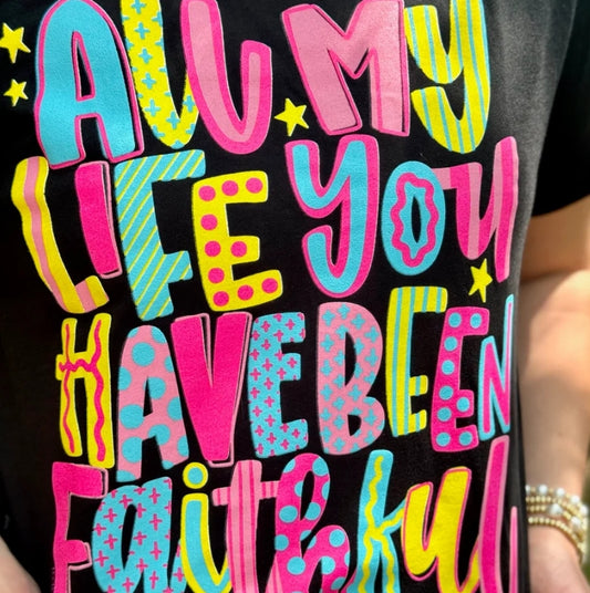 A close-up of a black T-shirt featuring a colorful graphic text that reads 'ALL MY LIFE YOU HAVE BEEN FAITHFUL' in playful, patterned letters with pink, blue, yellow, and green tones.