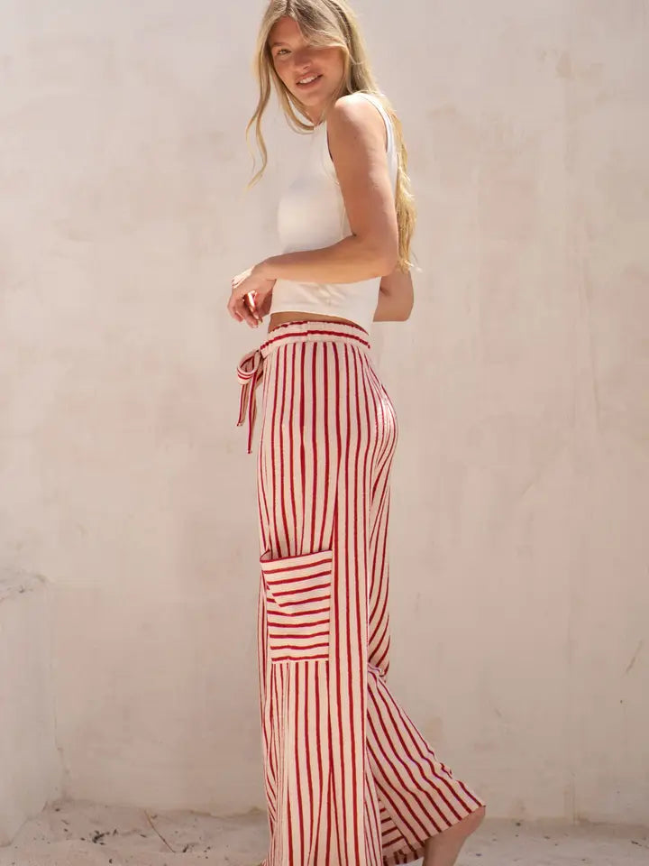 A woman wearing red and white vertically striped wide-leg pants with a matching tie belt, paired with a white sleeveless crop top, standing against a textured beige plaster wall.