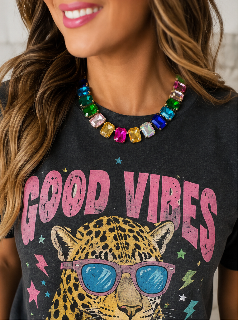A close-up lifestyle shot of a woman wearing a colorful gemstone rhinestone collar necklace and a black graphic t-shirt with a leopard print and pink 'GOOD VIBES' text.