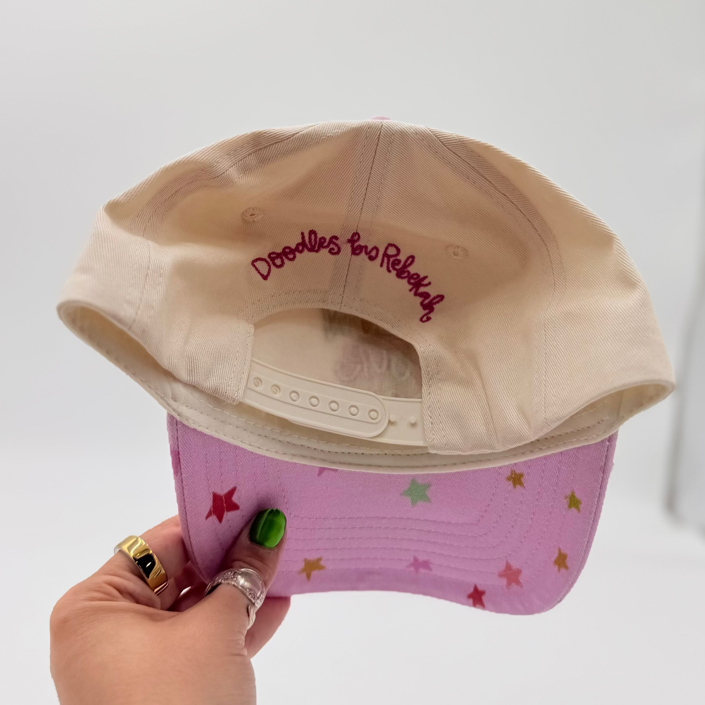 Beige and pink cap with 'Doodles are Rebels' text held by a hand on a white background