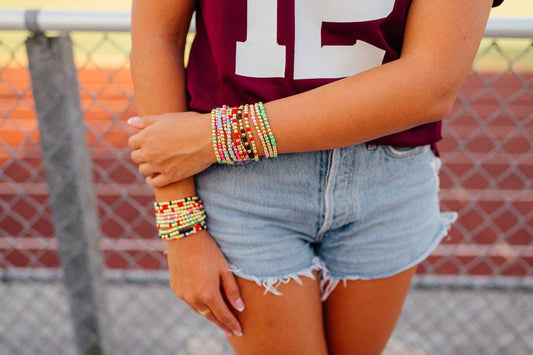 A person outdoors wearing multiple colorful beaded bracelets on both wrists, paired with a burgundy athletic top and light blue denim shorts.