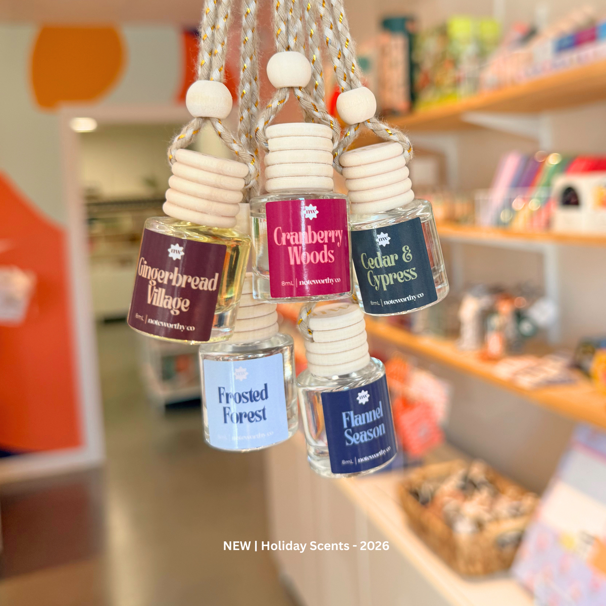 Five hanging glass perfume bottles with white wooden caps and cords, each labeled with a holiday scent name: Gingerbread Village, Cranberry Woods, Cedar & Cypress, Frosted Forest, and Flannel Season. The bottles are photographed in a softly blurred shop interior.