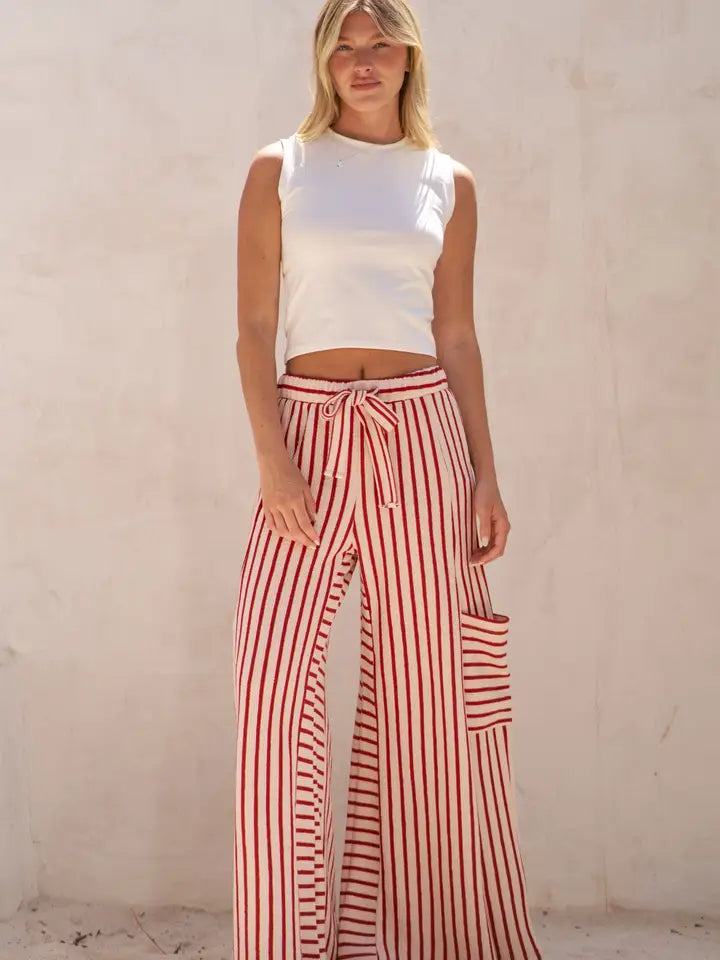 A woman modeling high-waisted, wide-leg pants with red and white vertical stripes, featuring a matching drawstring waist and a side cargo pocket, paired with a white sleeveless crop top against a textured beige wall.