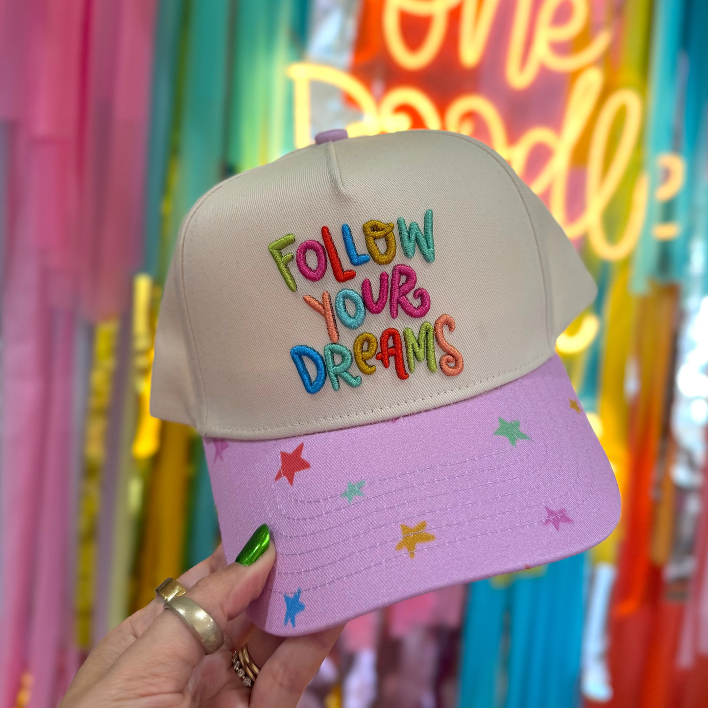 Colorful cap with 'Follow Your Dreams' text held in front of a vibrant, multicolored background.